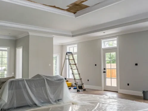 Ceiling water damage in a room with a ladder and furniture covered for repair.